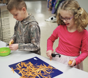 A boy and a girl using a peeler to cut carrots